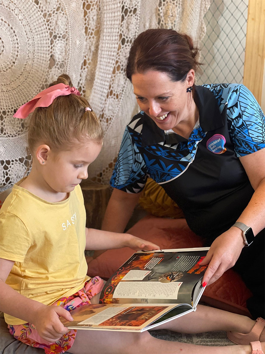 A child care worker and toddler sitting down and reading a book together.