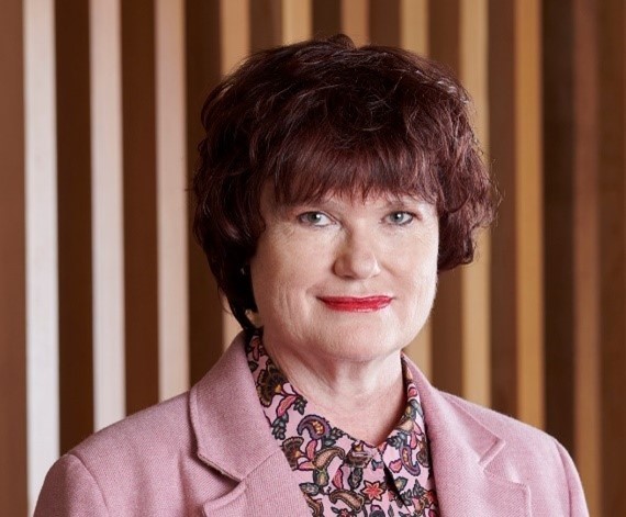 Headshot of Adrienne Nieuwenhuis - smiling woman with short reddish-brown hair, wearing reb lipstick, a pink floral shirt and light pink business jacket.