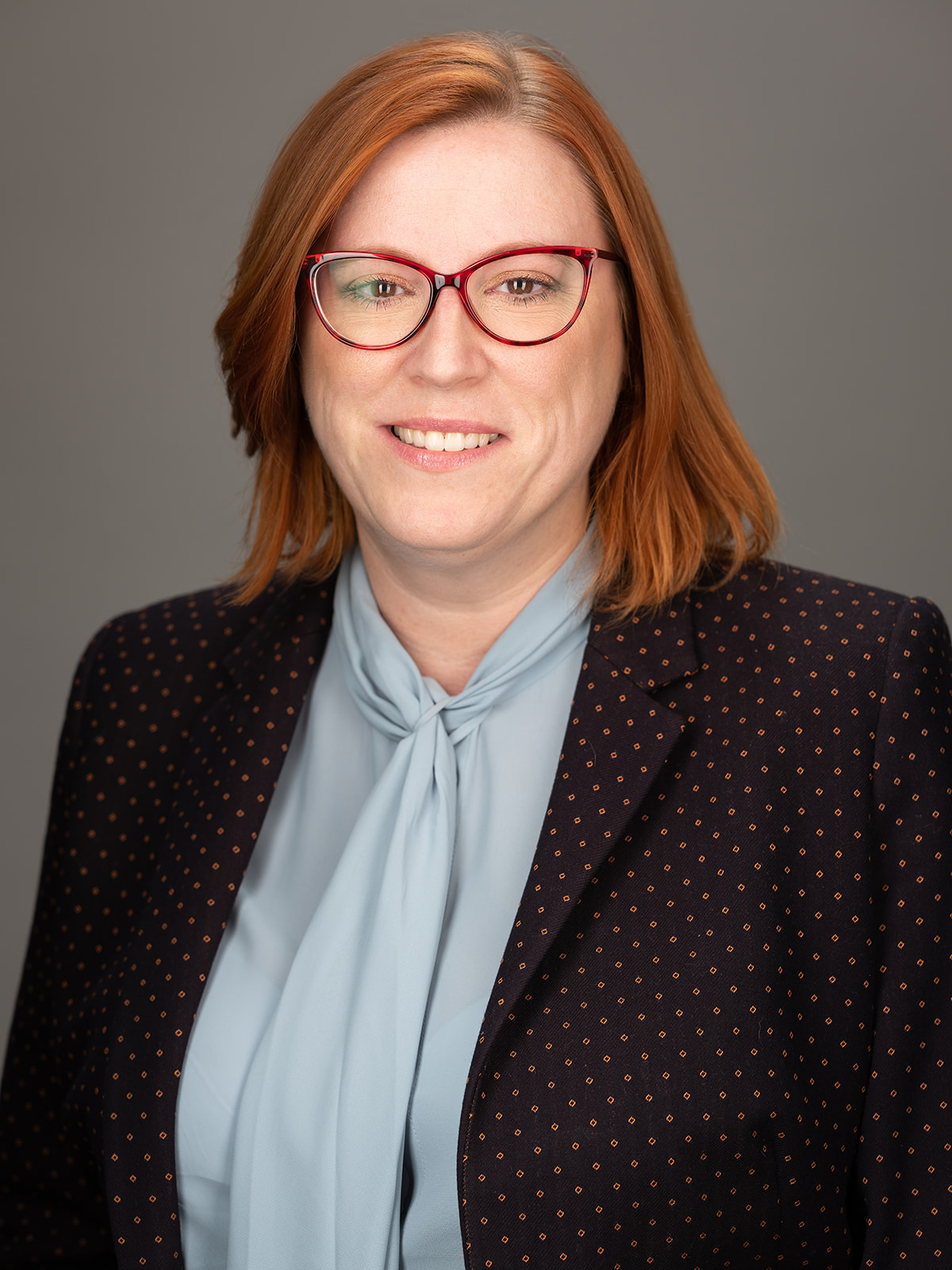 Headshot of Chloe Read - smiling woman with straight shoulder length ginger hair, wearing red glasses, a black business jacket and grey business shirt.