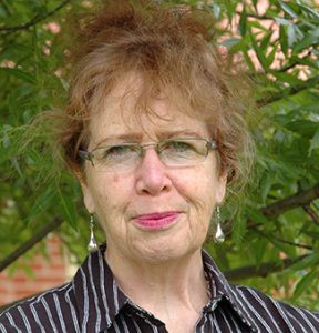 Headshot of Emeritus Professor Jo-Anne Reid - woman smiling with short light brown hair, wearing pink lipstick, silver earrings and glasses and a black coloured shirt with white stipes.